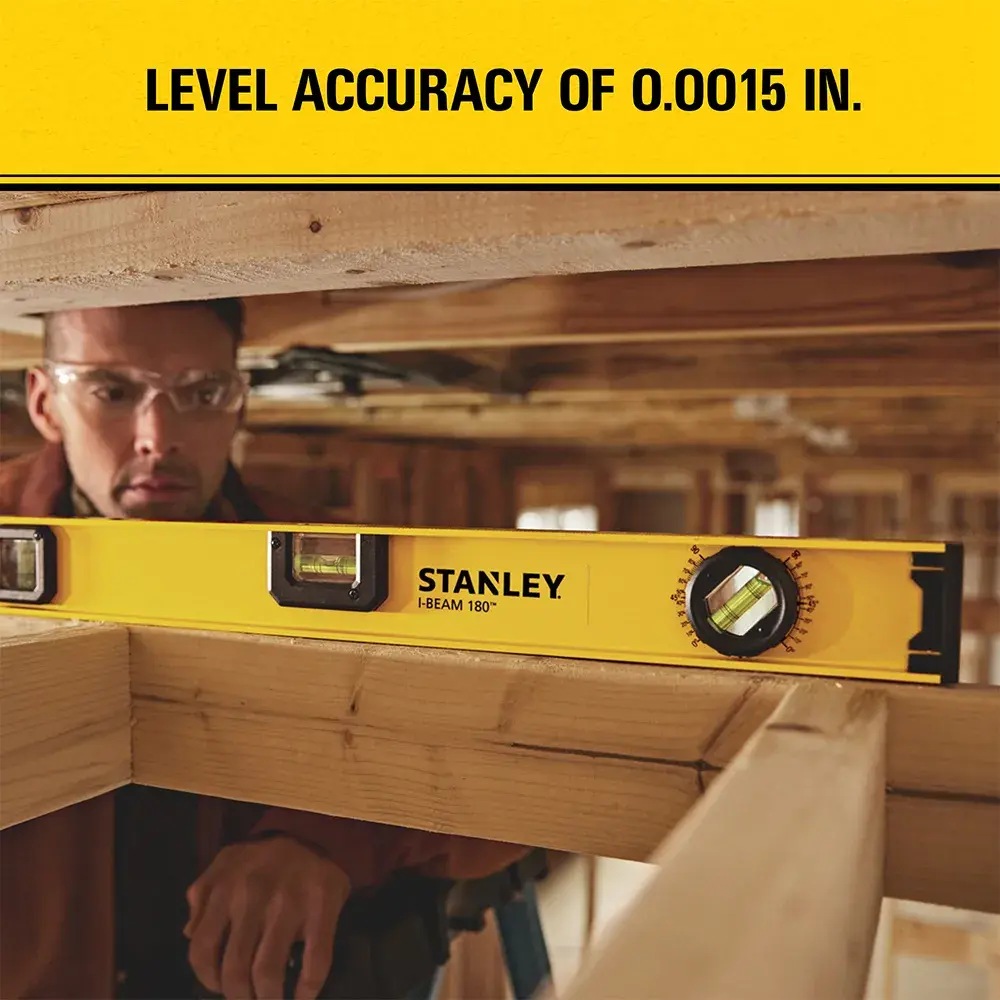 A yellow Stanley spirit level (model 42-324) is placed on wooden beams in a construction setting. The spirit level displays its brand name and features high accuracy of 0.0015 inches, as stated in the upper part of the image. A person is working behind the beams, with their face blurred out.