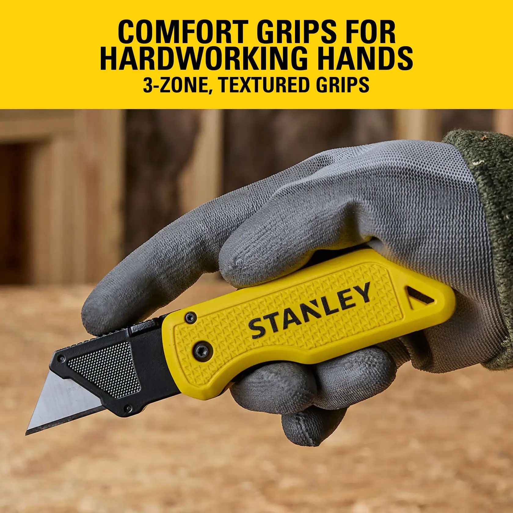 Gloved hand holding a yellow Stanley utility knife with textured grip and sharp blade on a wood surface.