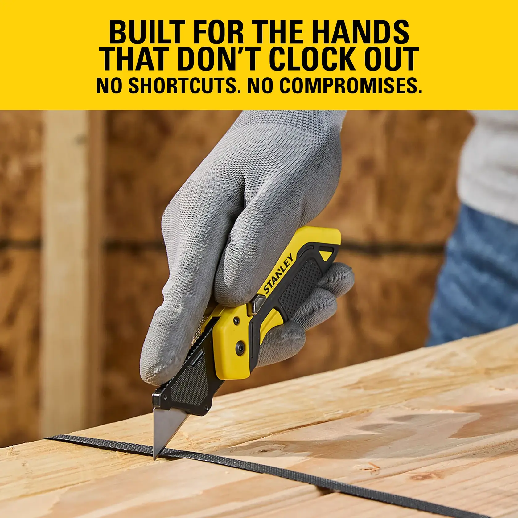 Stanley utility knife held by gloved hand, cutting material on wooden surface in a workshop setting.