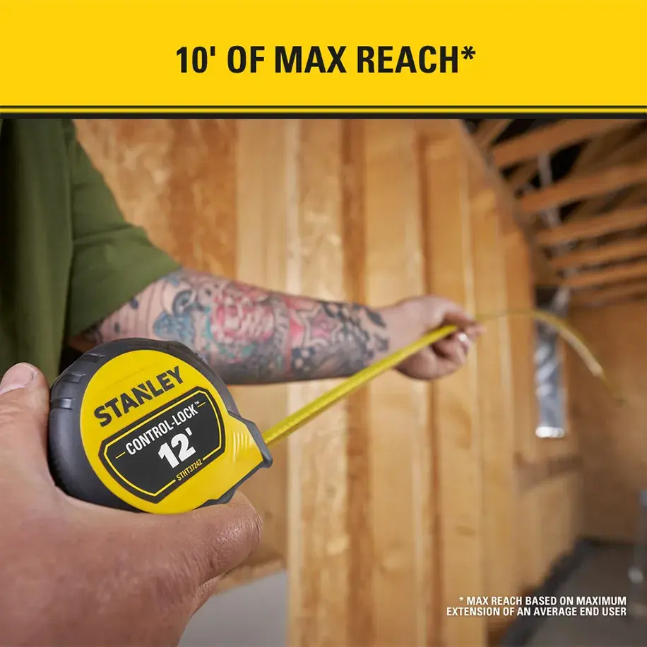 A person uses a Stanley Control-Lock 12 tape measure inside a wooden framed construction area. The yellow tape is extended, and the text at the top reads '10' OF MAX REACH'.