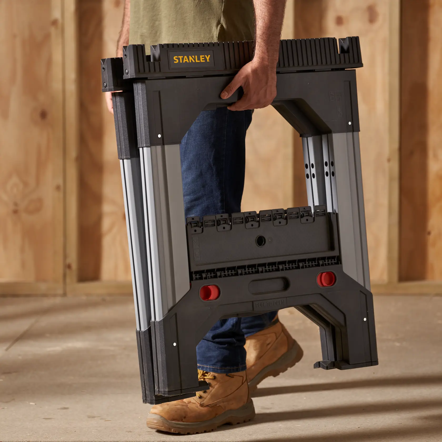 Stanley 011031S folding sawhorse, black and silver, carried by person in work boots, workshop setting.