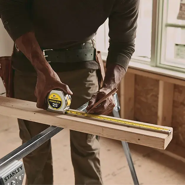 A person using a Stanley tape measure to mark a piece of wood inside a construction or carpentry workspace.