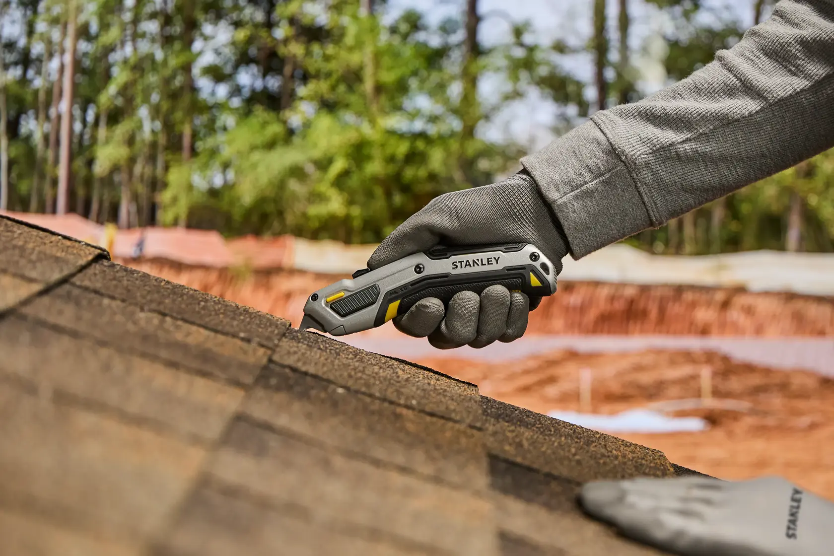 A person wearing gray gloves uses a Stanley utility knife to cut roofing shingles outdoors.