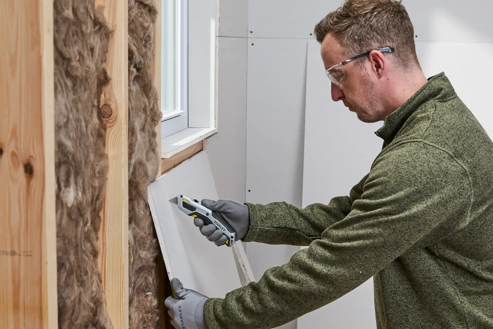 A person wearing gloves uses a STANLEY utility knife to cut drywall near a window, next to insulation and wooden wall studs.