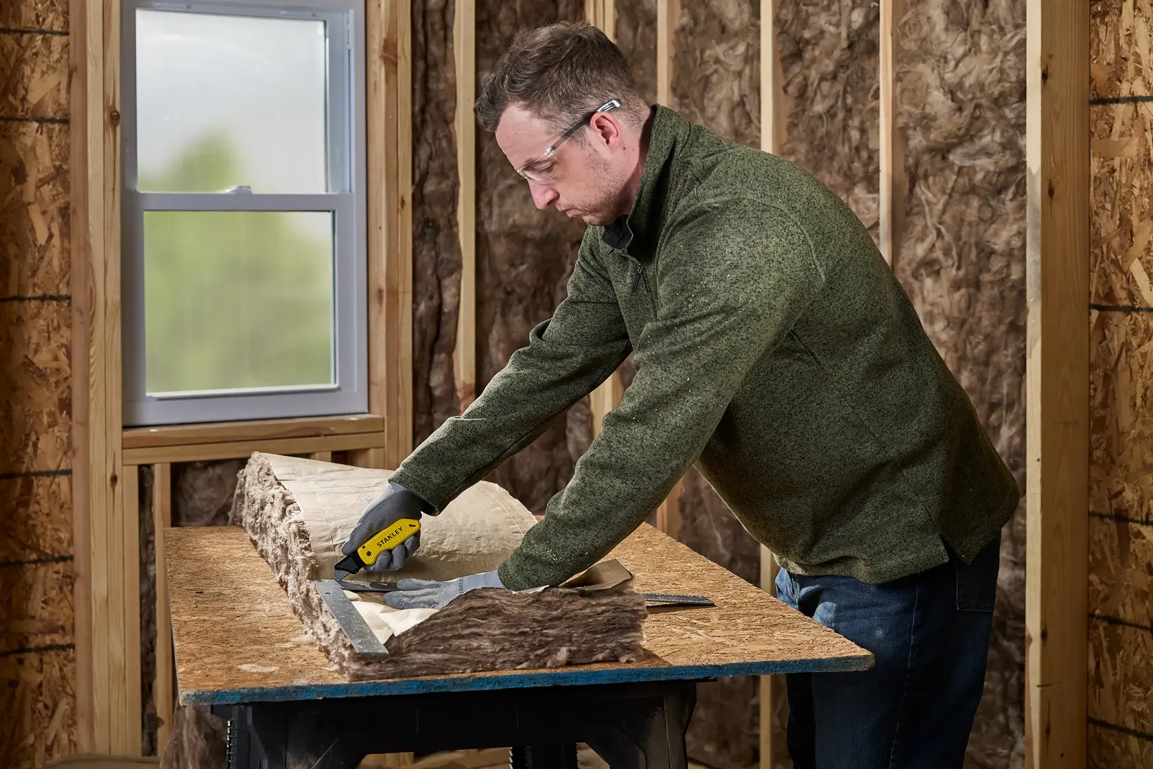 A person wearing gloves uses a STANLEY utility knife to cut insulation material on a workbench in a room under construction.