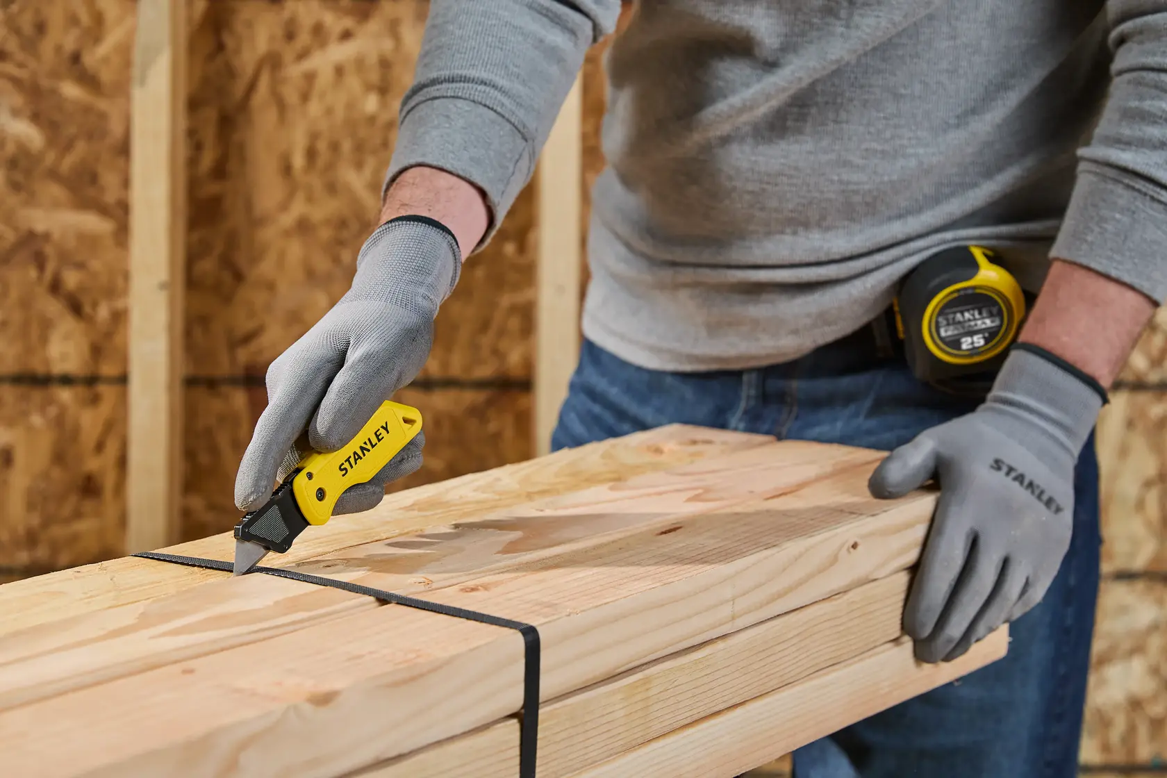 A person wearing gray gloves uses a yellow STANLEY utility knife to cut black straps securing a bundle of wooden planks. A STANLEY tape measure is attached to their belt. The scene appears to be in a workshop or construction setting.