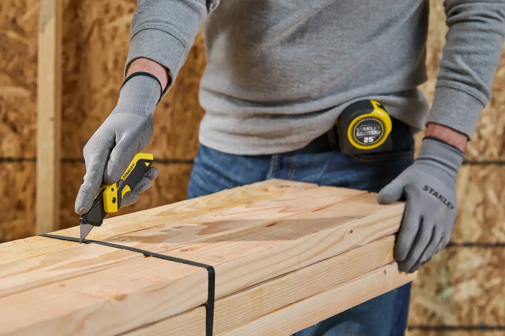 A person wearing Stanley branded gloves uses a Stanley utility knife to cut a black strap on a stack of wooden beams. The person also has a Stanley tape measure attached to their belt.