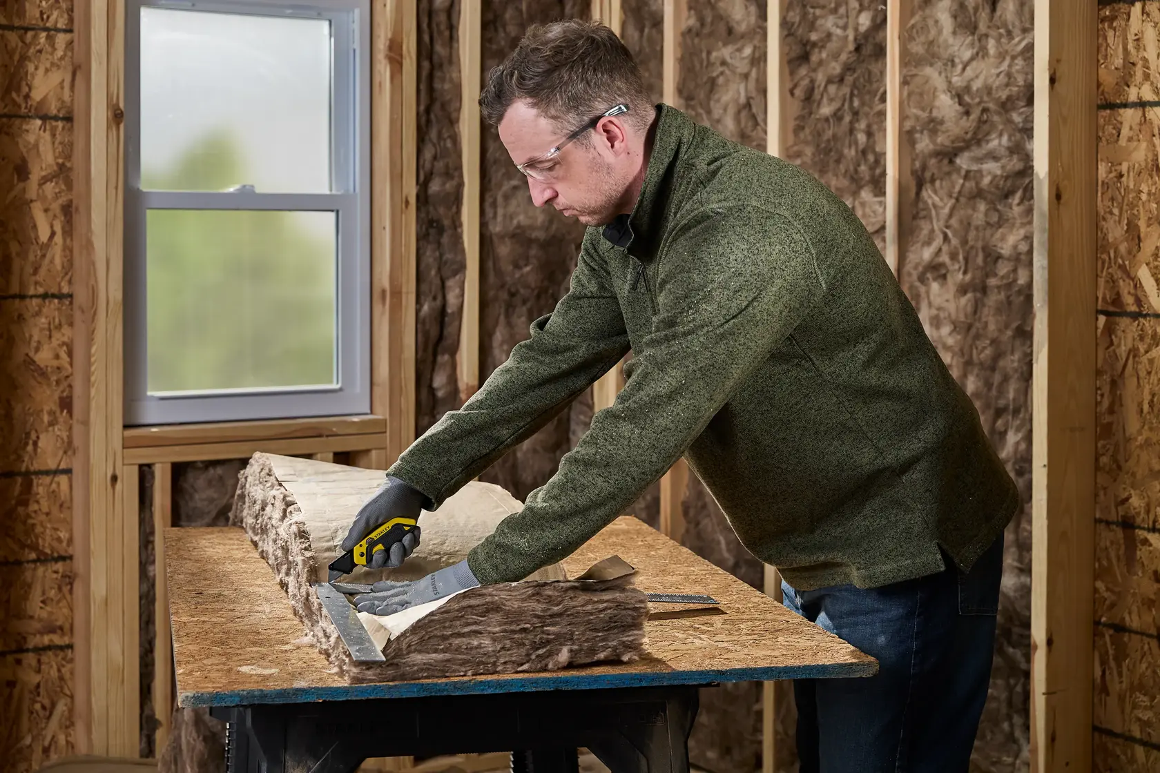 A person wearing gloves uses a Stanley utility knife to cut insulation material on a workbench inside a room under construction with exposed wooden framing.