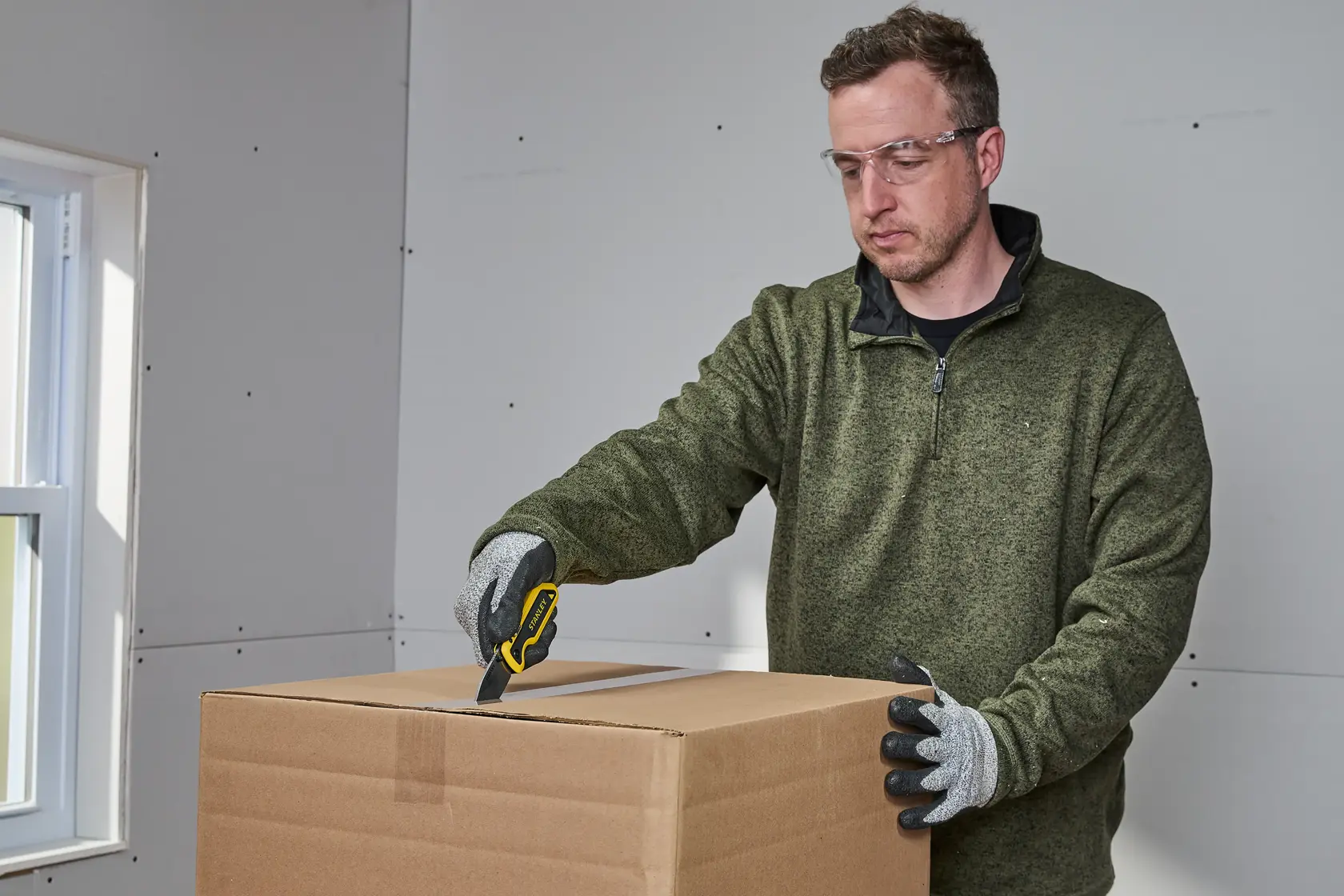 Person wearing gloves uses a STANLEY utility knife to cut open a cardboard box in an indoor setting. The person's face is blurred.