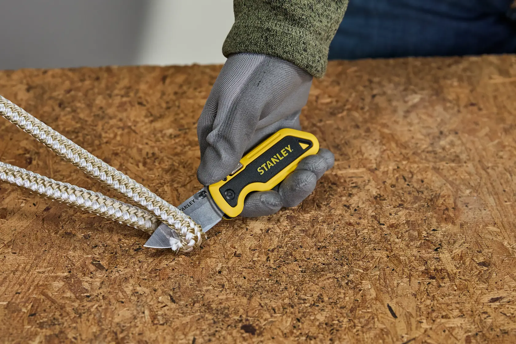 A gloved hand using a STANLEY utility knife to cut a braided rope on a wooden surface.