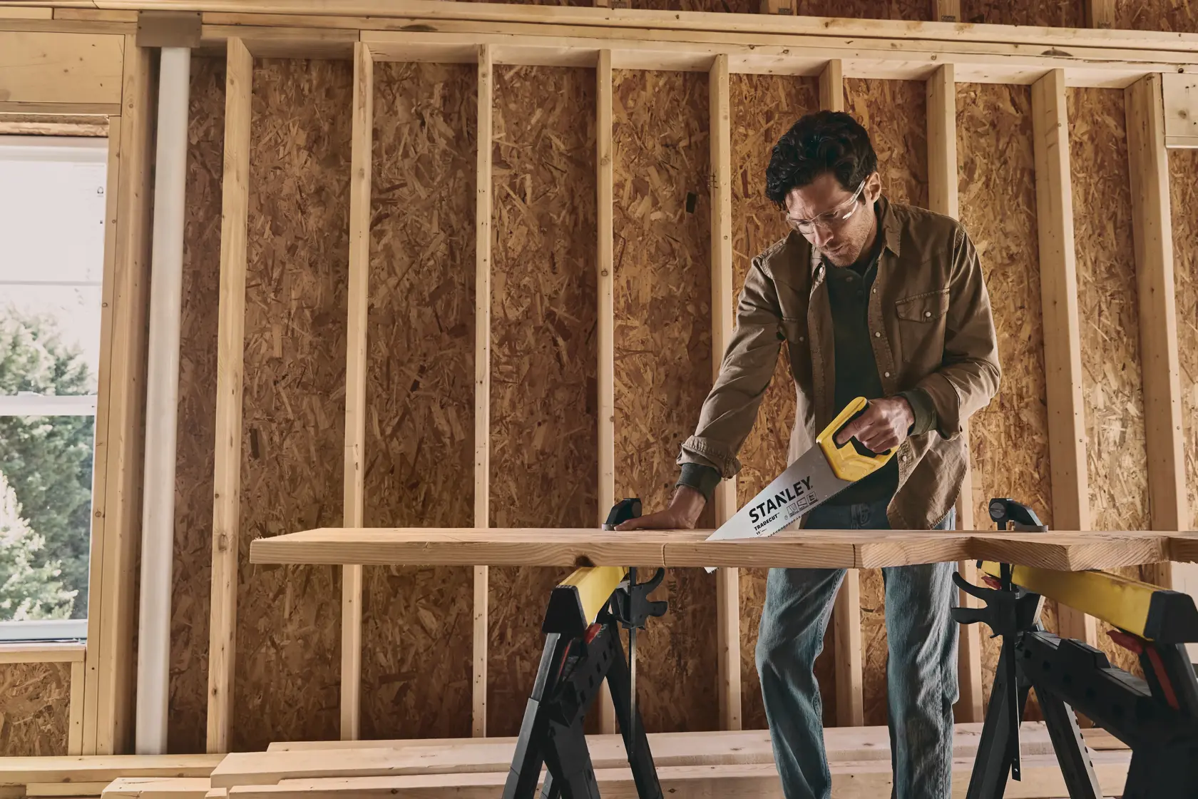 STANLEY STHT20348 hand saw used to cut wood plank on sawhorses inside unfinished building.