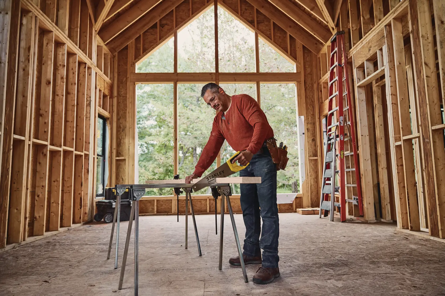 STANLEY STHT20348 hand saw used to cut wood plank on sawhorses inside a wooden house frame under construction.