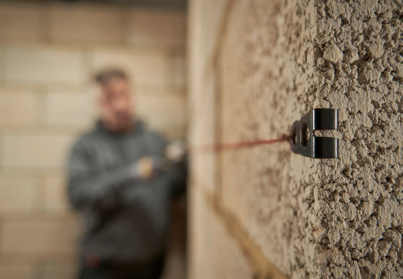 A close-up view of a masonry wall with a STANLEY line block attached and a taut red line running along the wall. In the background, a person in a grey hoodie is blurred, setting the line for construction alignment.