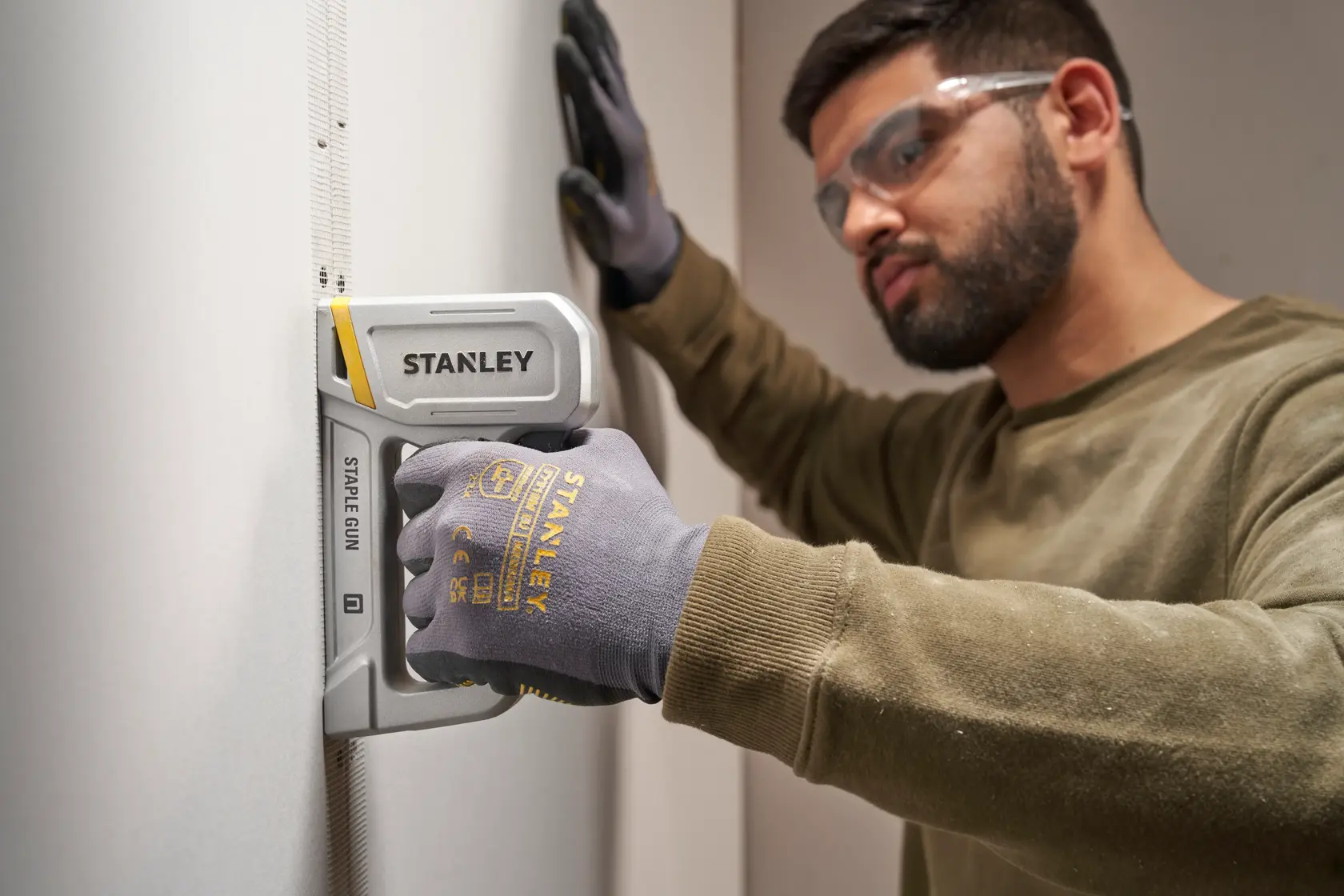 Photo of STANLEY STHT70150 staple gun in use, held by a gloved hand against a wall, side angle.