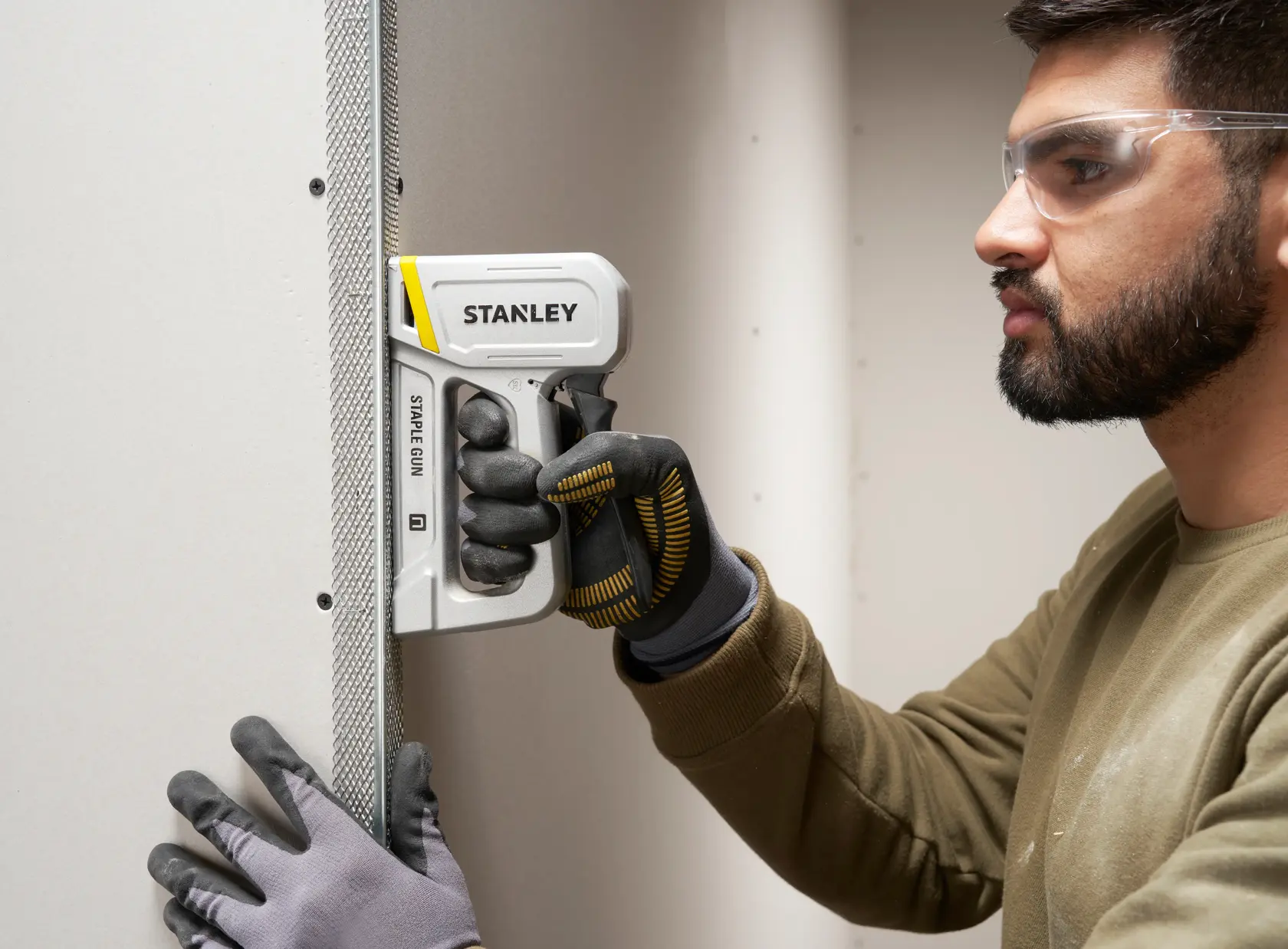 STANLEY STHT70150 staple gun used on drywall with gloved hands, close-up photography.