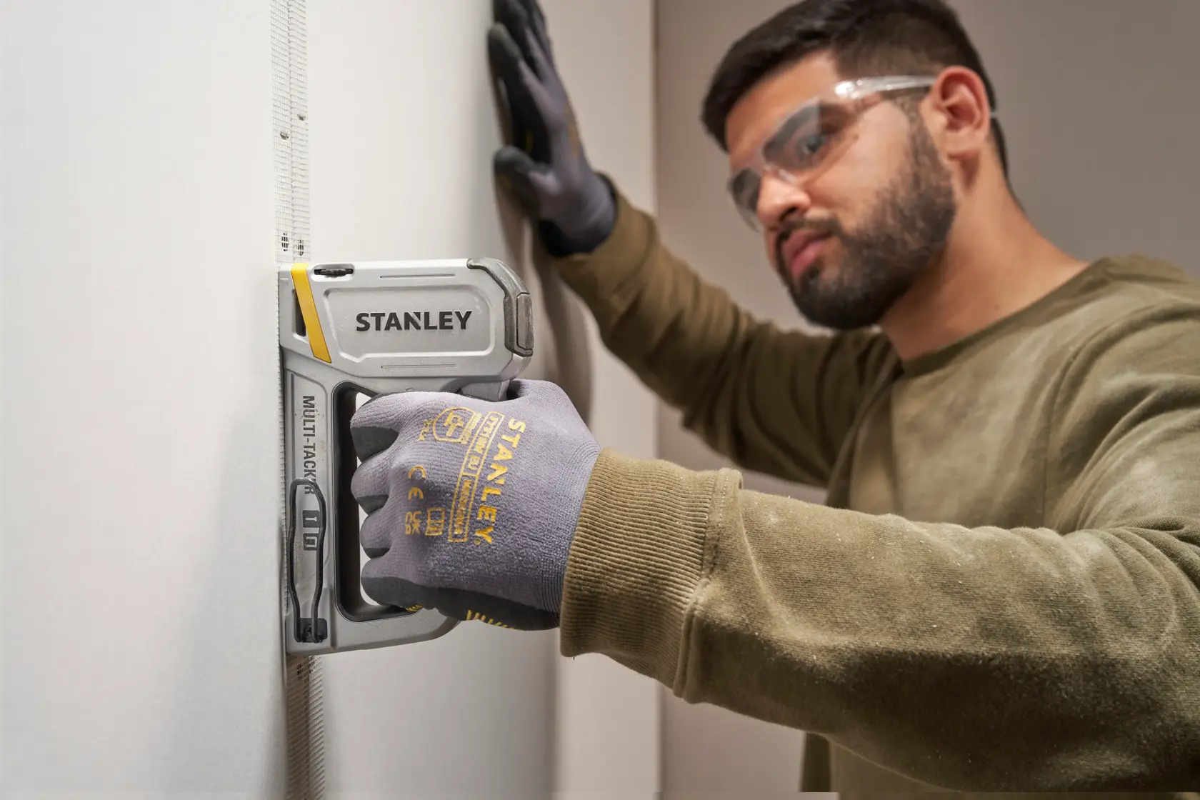 Photo of a person wearing gloves using a STANLEY STHT70250 stapler on drywall, close-up view.