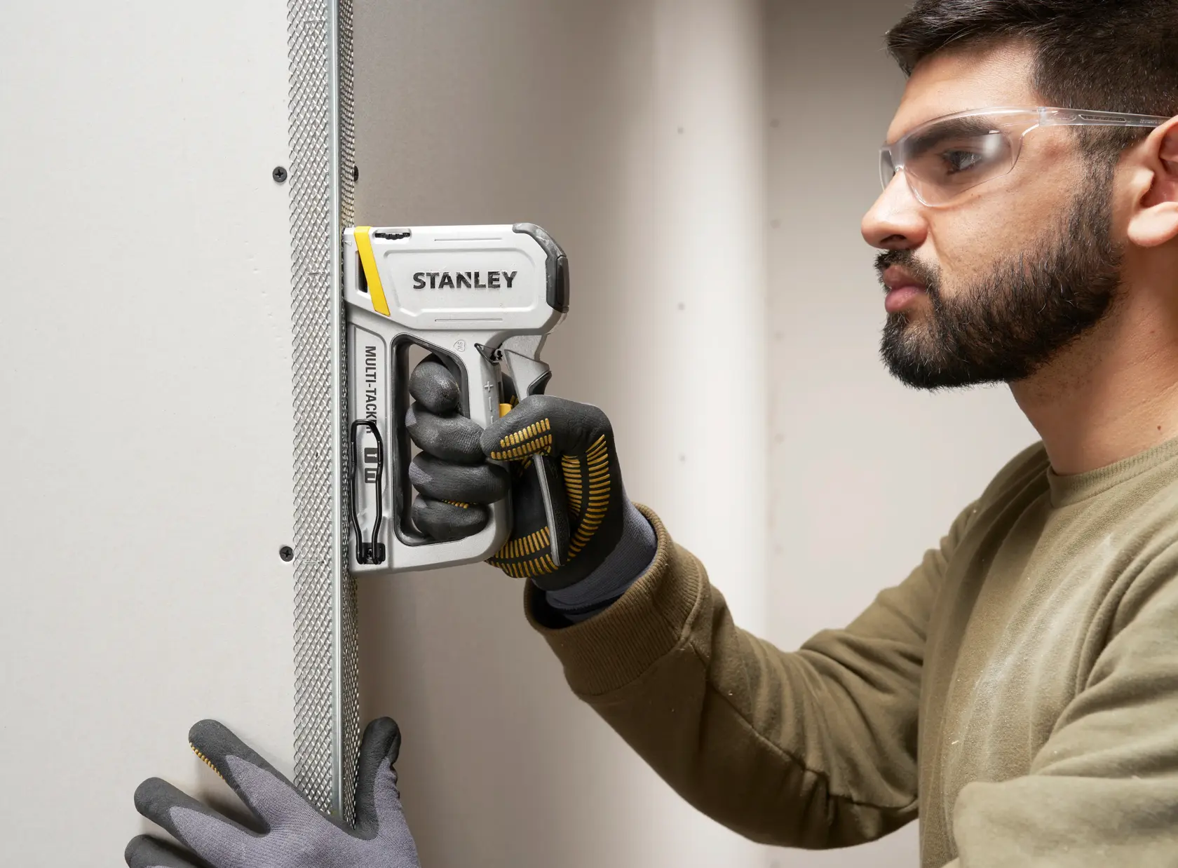 Photo of STANLEY STHT70250 staple gun in use, held by gloved hands on drywall and metal corner bead.