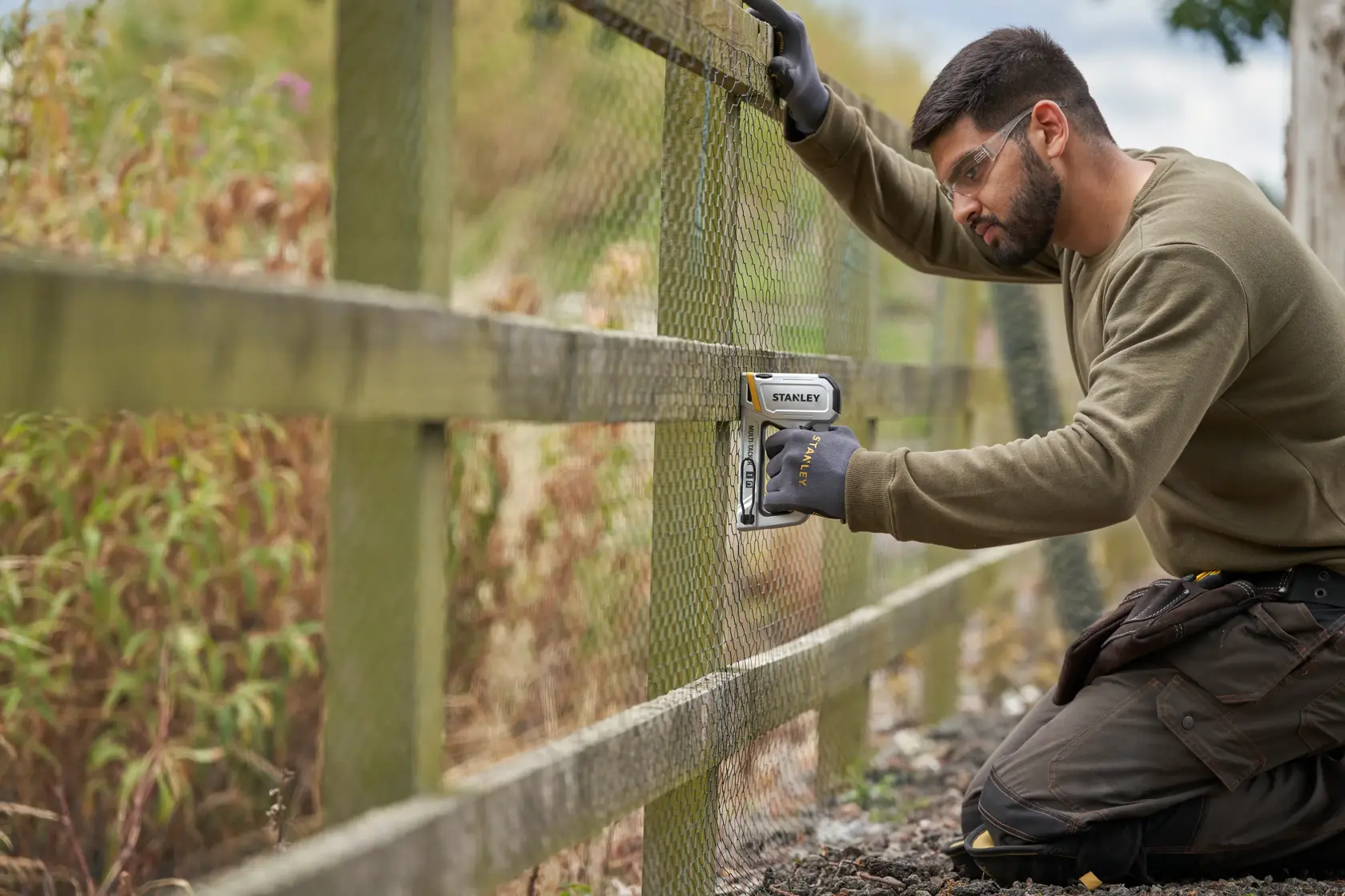 STANLEY STHT70250 staple gun used on an outdoor wooden fence to secure wire mesh, person wearing work gloves.