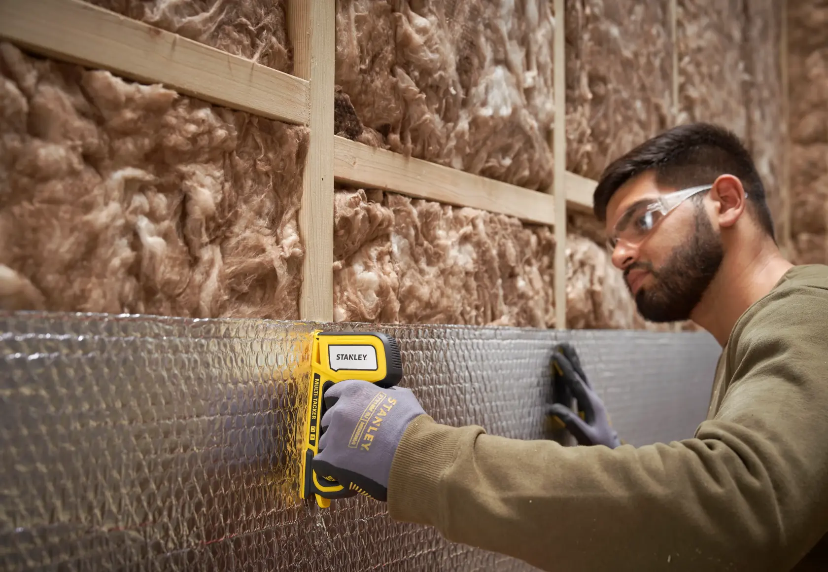Stanley STHT70440 hand stapler used to fasten insulation material to a wall frame, person wearing work gloves.
