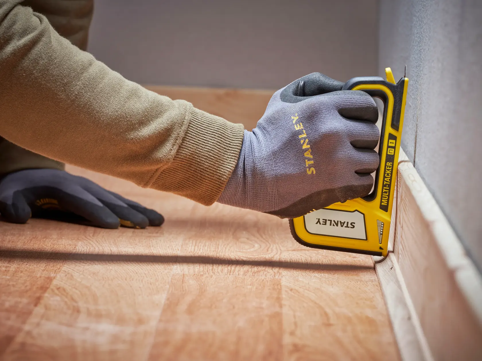 Photo of STANLEY STHT70440 manual stapler being used on floor trim, close-up of gloved hand holding tool.