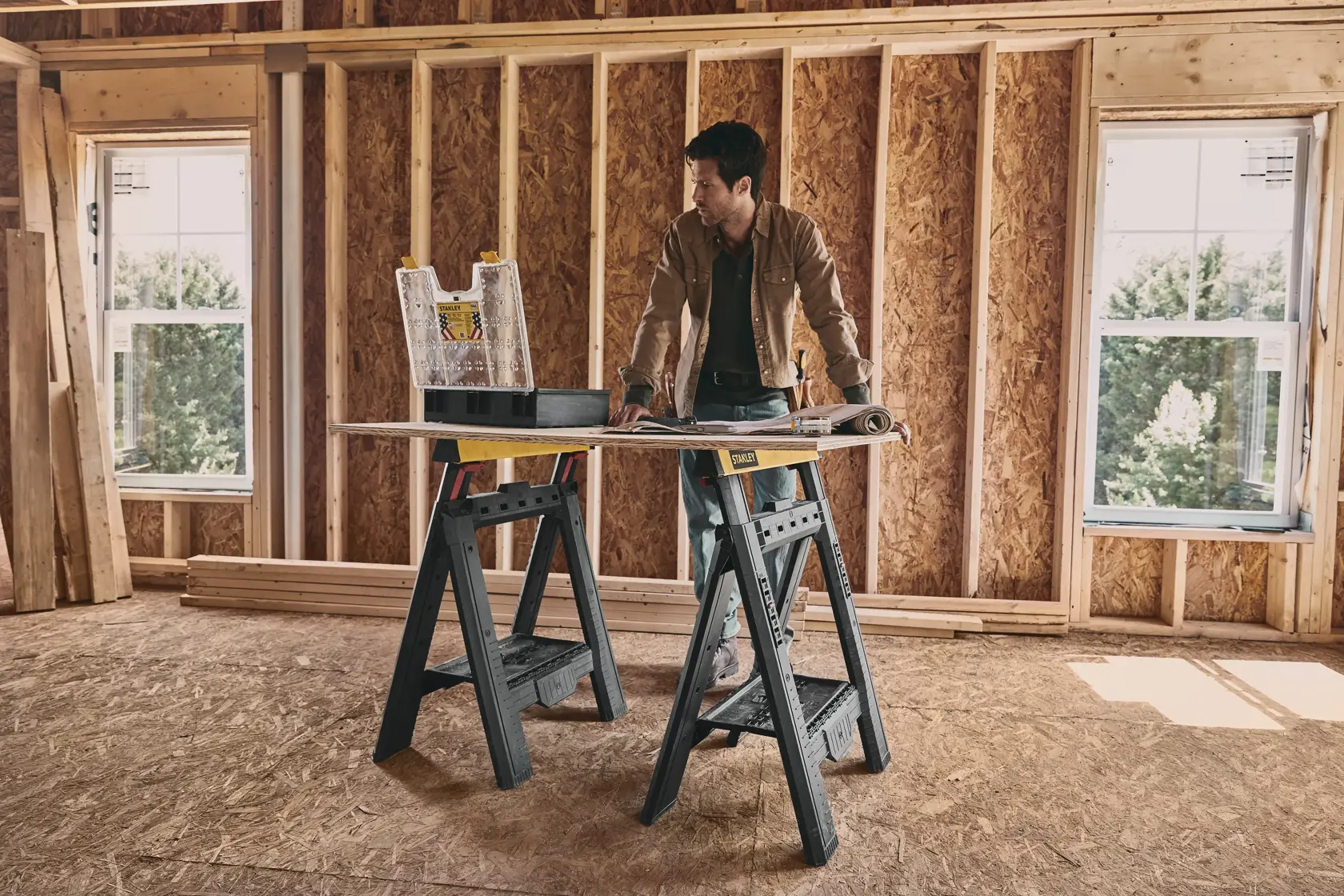 STANLEY STST60626 sawhorse set supports a work surface in a construction room, with tools and plans on top.
