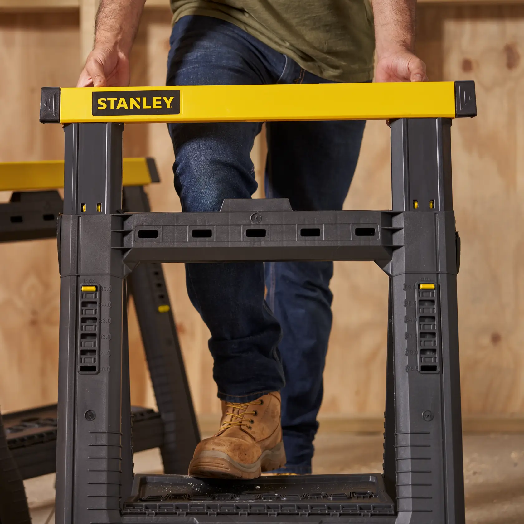 STANLEY STST60626 plastic adjustable sawhorse, front angle, yellow and black, workshop setting.