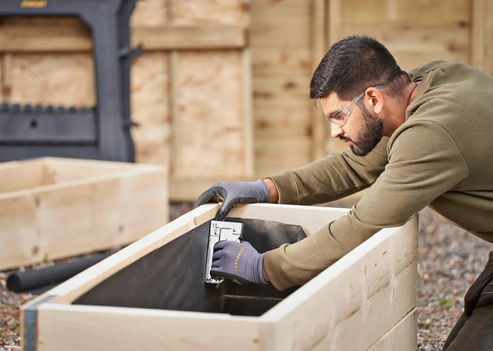 STANLEY TR45 stapler used to secure fabric inside a wooden garden bed, user wearing gloves outdoors.